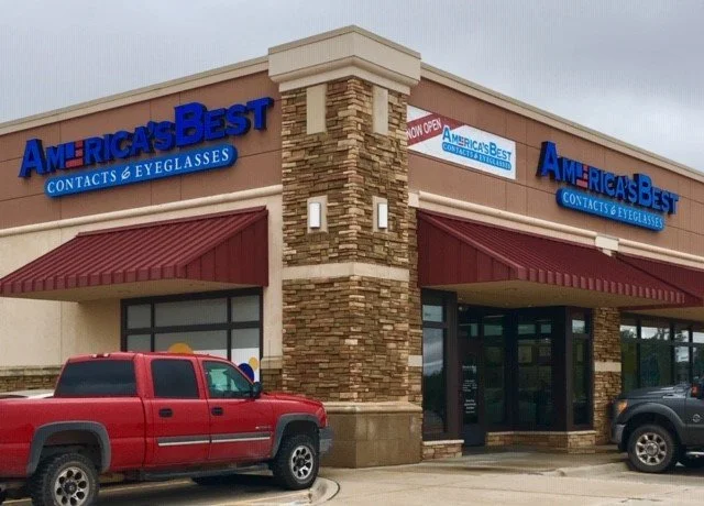 America's Best Contacts & Eyeglasses store in a shopping plaza with a brick and beige exterior, red awnings, and parked vehicles in front.
