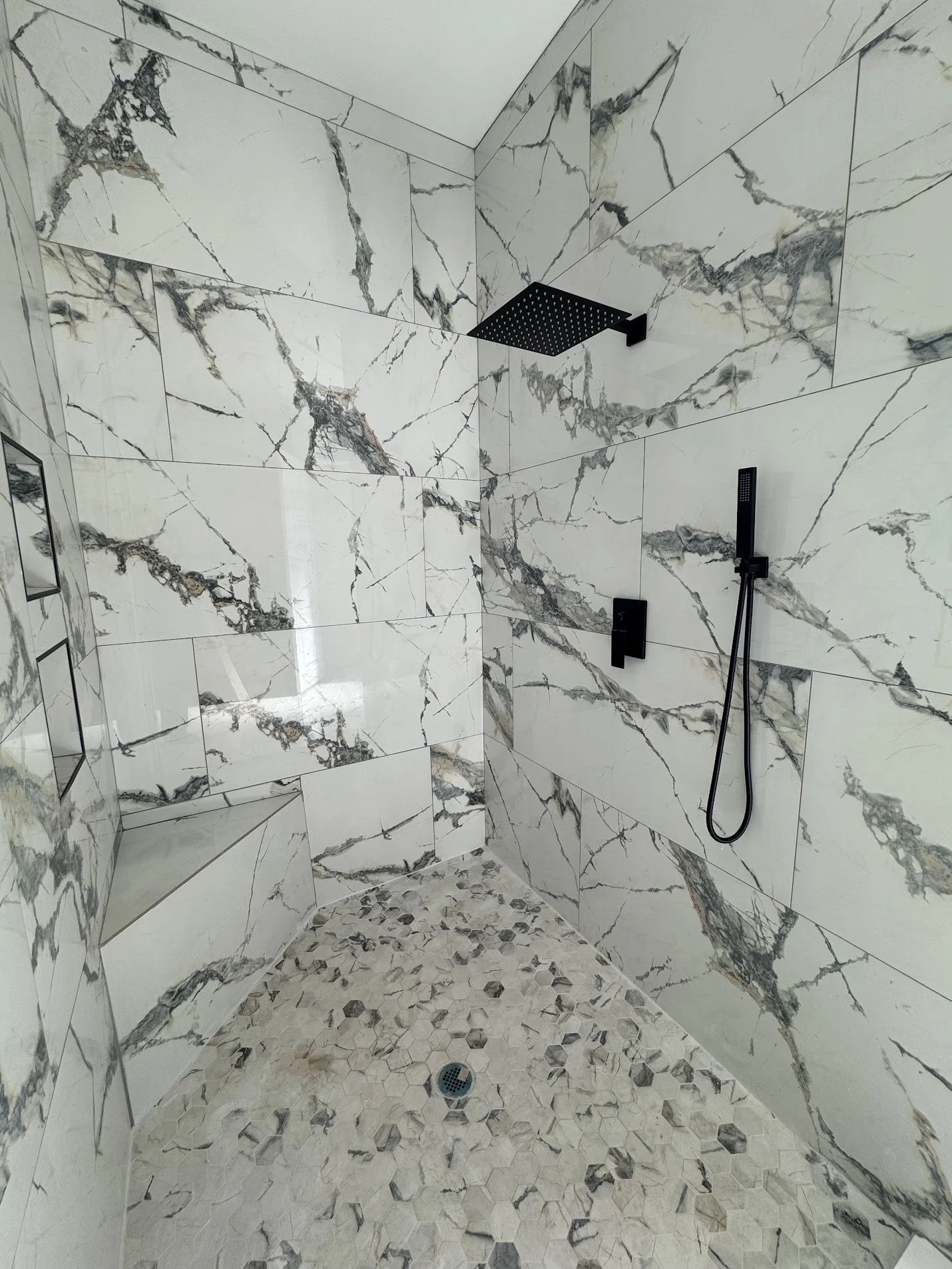 Modern shower with white marble tiles with dark gray and brown veining, black shower fixtures, and a pebble tile floor.