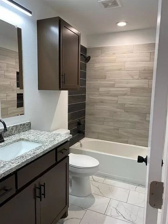 Modern small bathroom with a granite countertop sink, dark brown cabinets, a toilet, a bathtub with a tiled surround, and a walk-in shower area behind a partial wall.