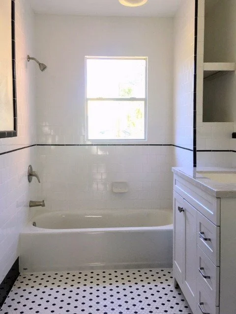 Bathroom with white tile walls, black trim, a bathtub, a window, a bathroom vanity with drawers, and black and white mosaic floor tiles.