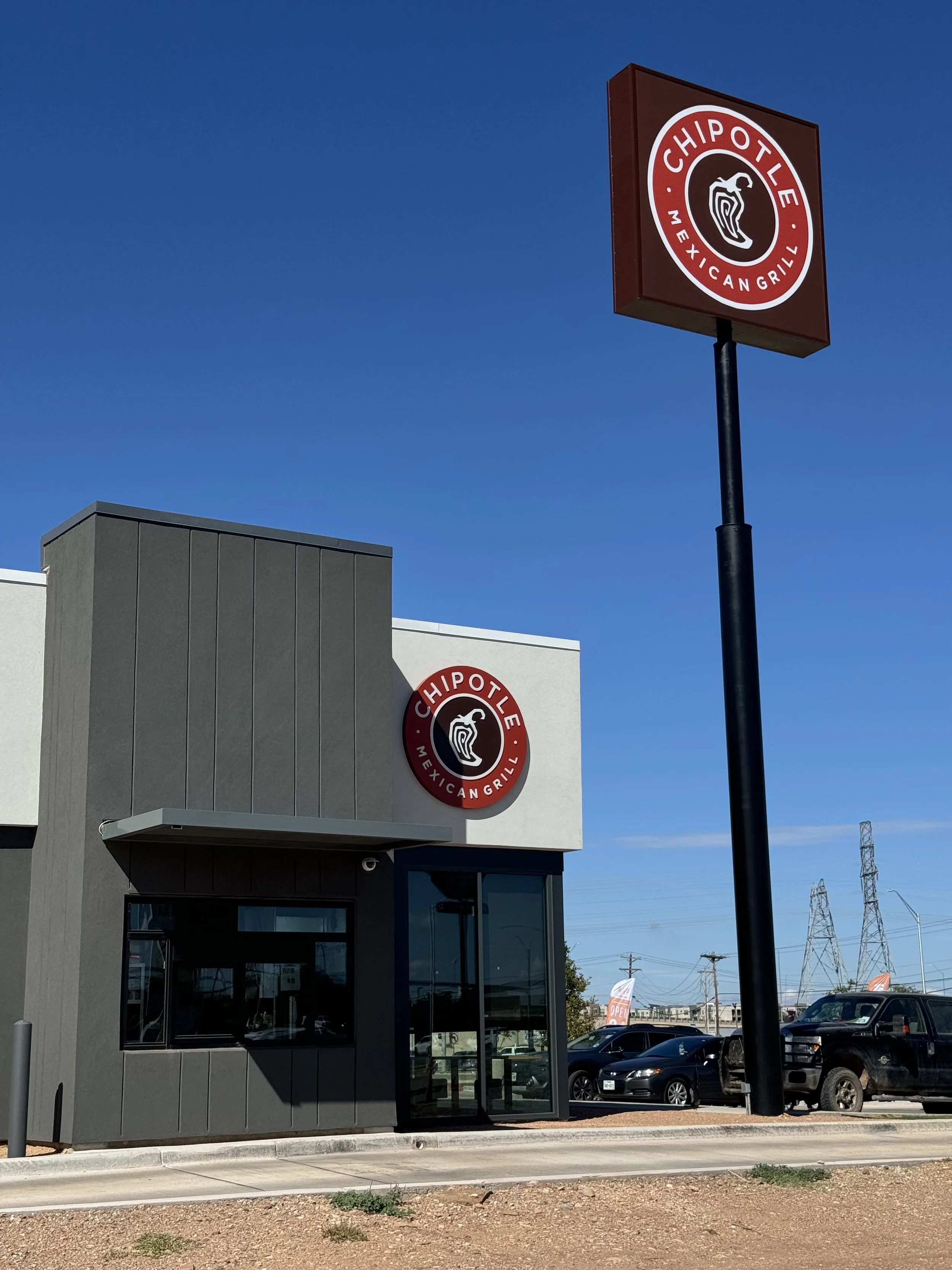 Exterior view of a Chipotle restaurant with the Chipotle logo on the building and a standalone sign under a clear blue sky.
