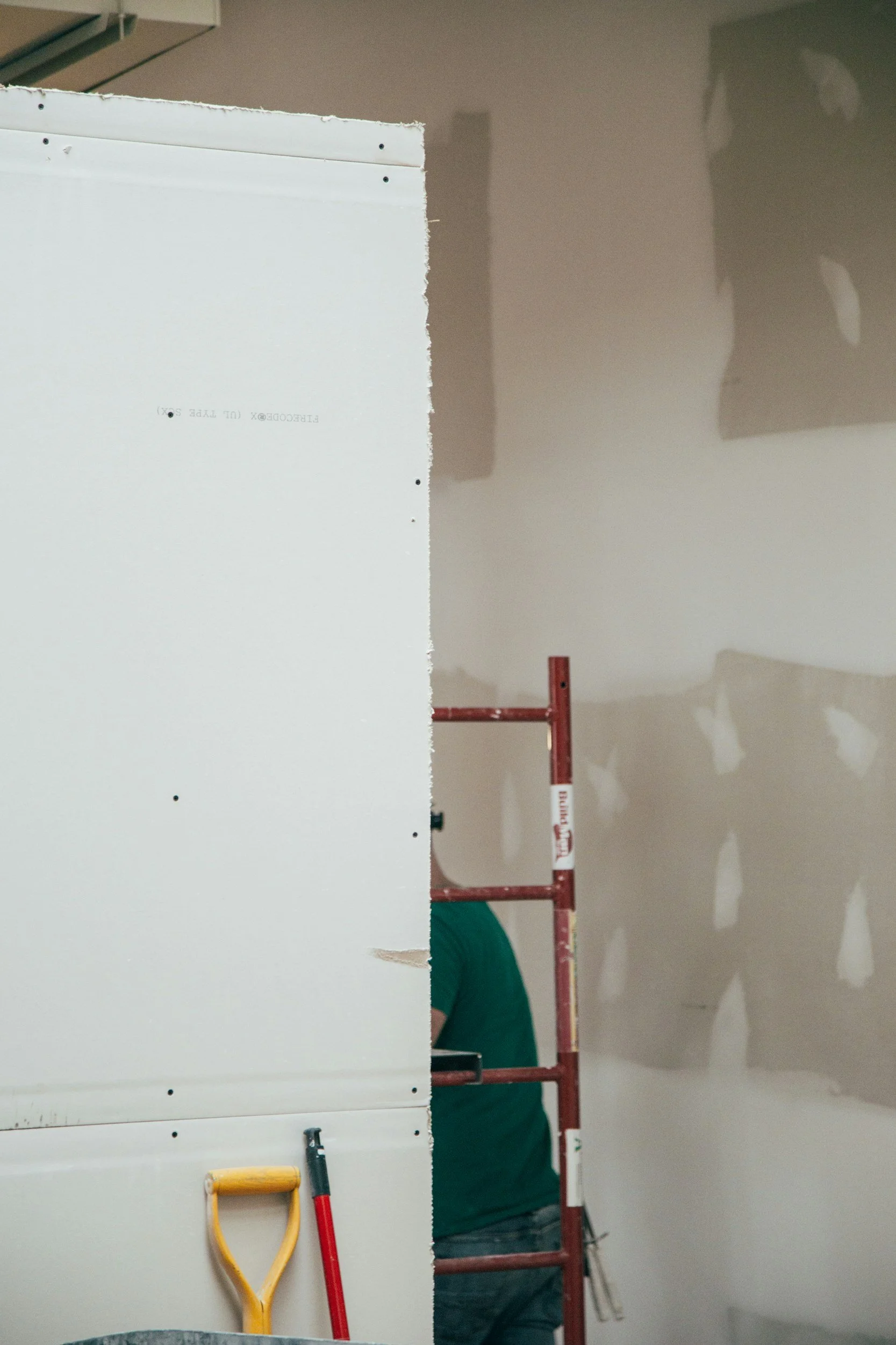 Partially finished drywall in a room under construction or renovation, with tools such as a yellow-handled trowel and a red-handled tool resting against it, and a worker in a green shirt standing on a scaffold in the background.