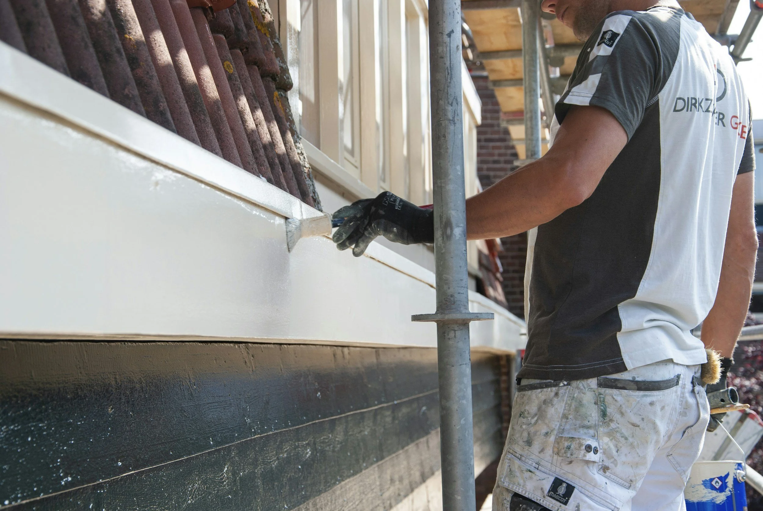 A worker painting the exterior of a house, applying white paint to the house's siding while standing on scaffolding.