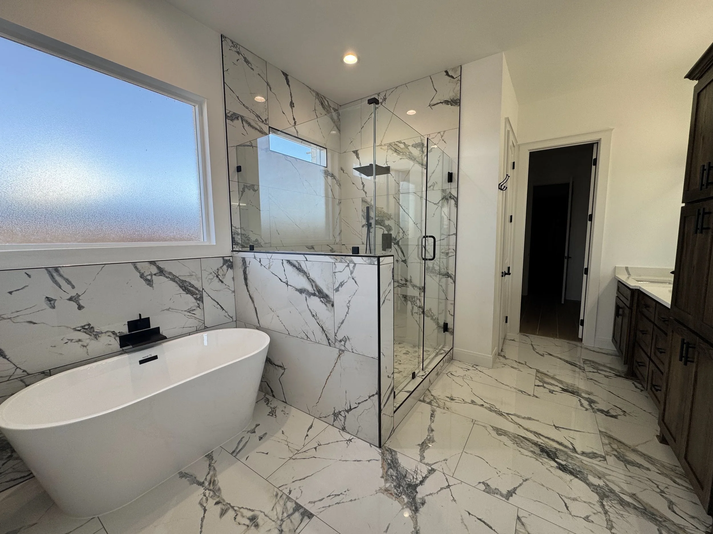 Modern bathroom with white and black marble tiles; freestanding bathtub, glass-enclosed shower, large frosted window, wooden cabinetry, and a white countertop.