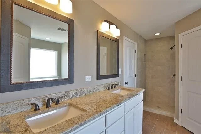 Bathroom with double sinks, granite countertop, large mirror, and beige tiled shower area.