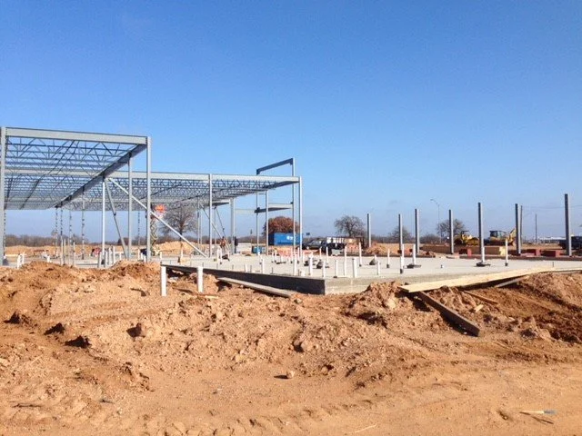 Construction site with a steel framework structure being built on a concrete foundation, dirt in the foreground, and a clear blue sky.