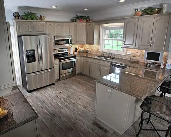 A spacious kitchen with gray cabinets, stainless steel appliances, granite countertops, a window above the sink, and a small TV on the countertop.