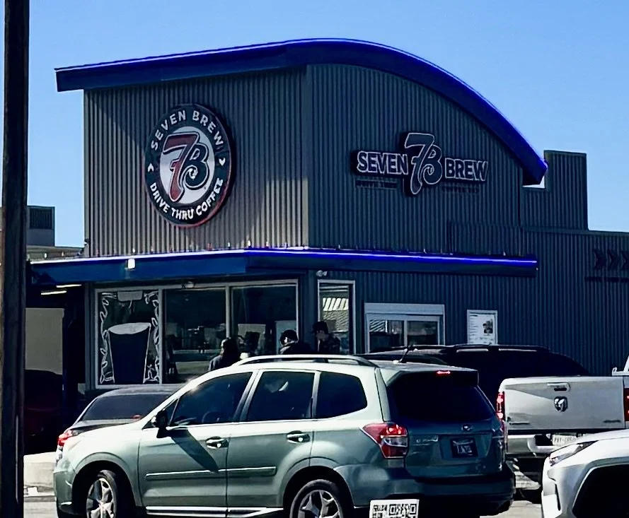 Exterior of Seven Brew drive-thru coffee shop with parked cars in front, people inside, and signage displaying the name.