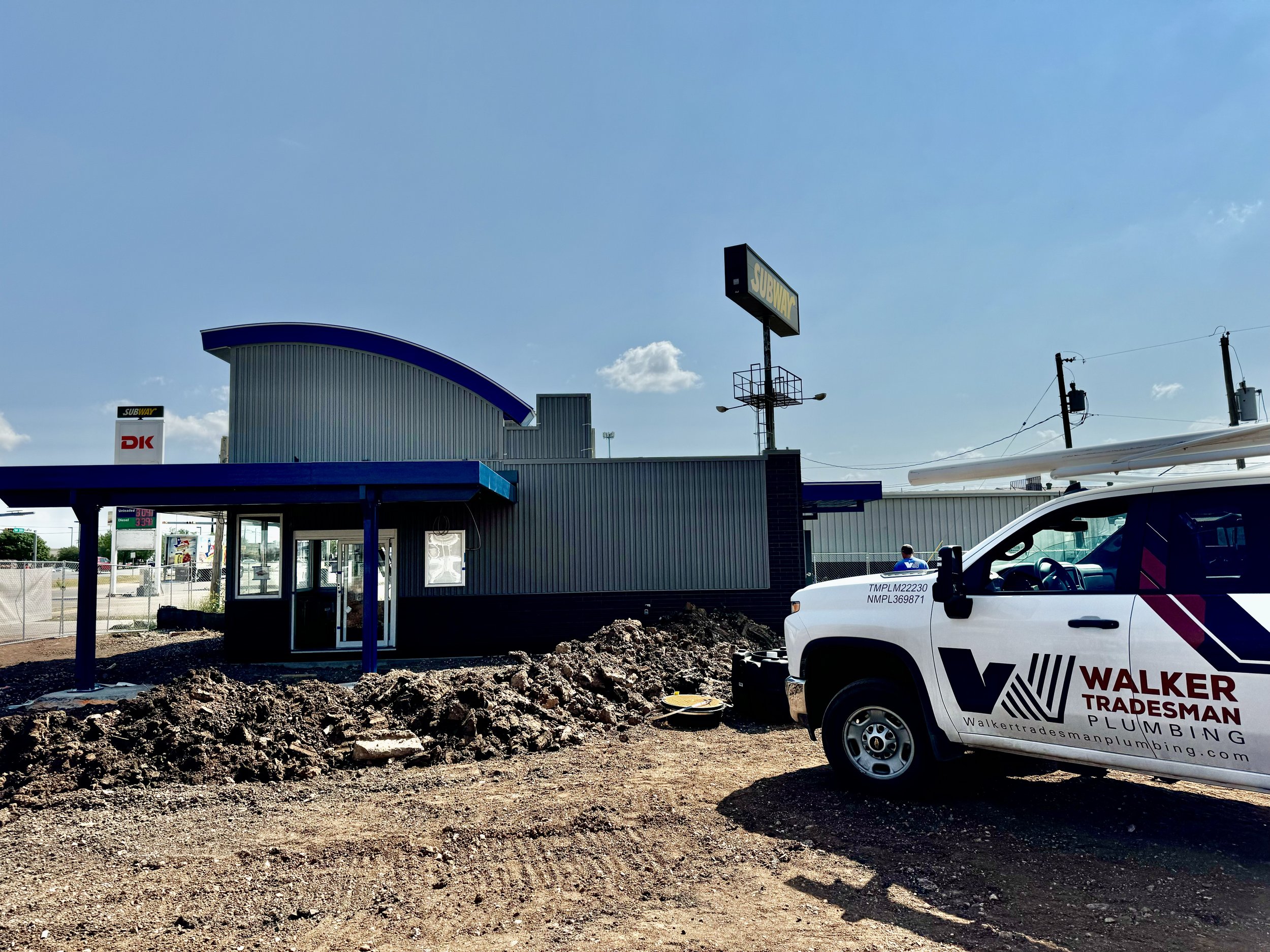 Construction site in front of a Subway restaurant with a white van bearing the Walker Tradesman Plumbing logo parked nearby. Dirt piles and construction equipment are visible.