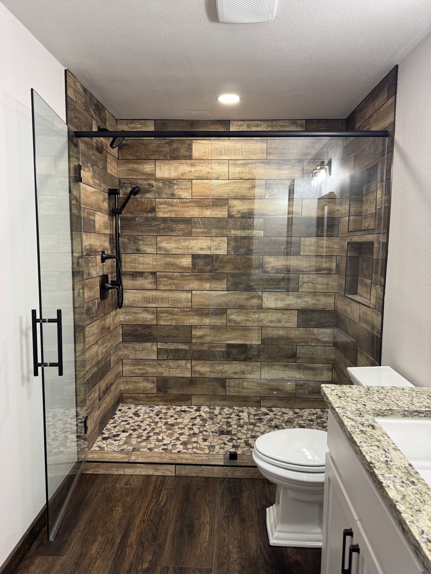 Modern bathroom featuring a walk-in shower with wood-look tile walls and pebble tile floor, a glass shower door, white vanity with granite countertop, and a toilet.