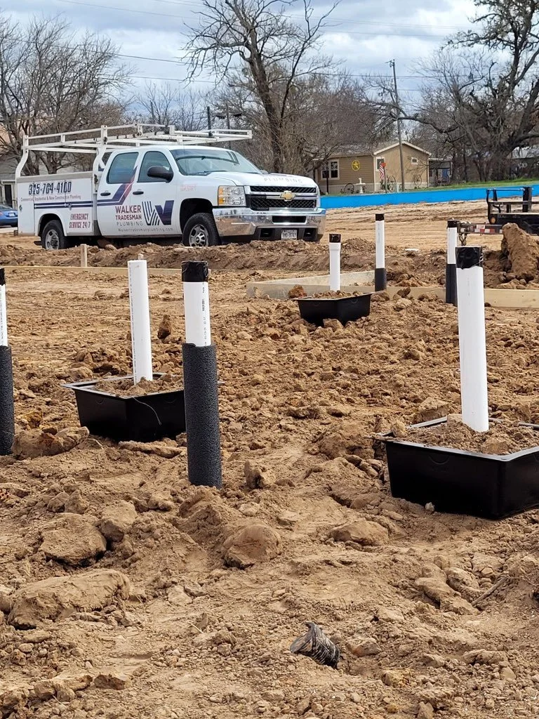 Construction site with white PVC pipes and black landscaping edging, a white truck parked in the background, and trees and a house visible beyond.