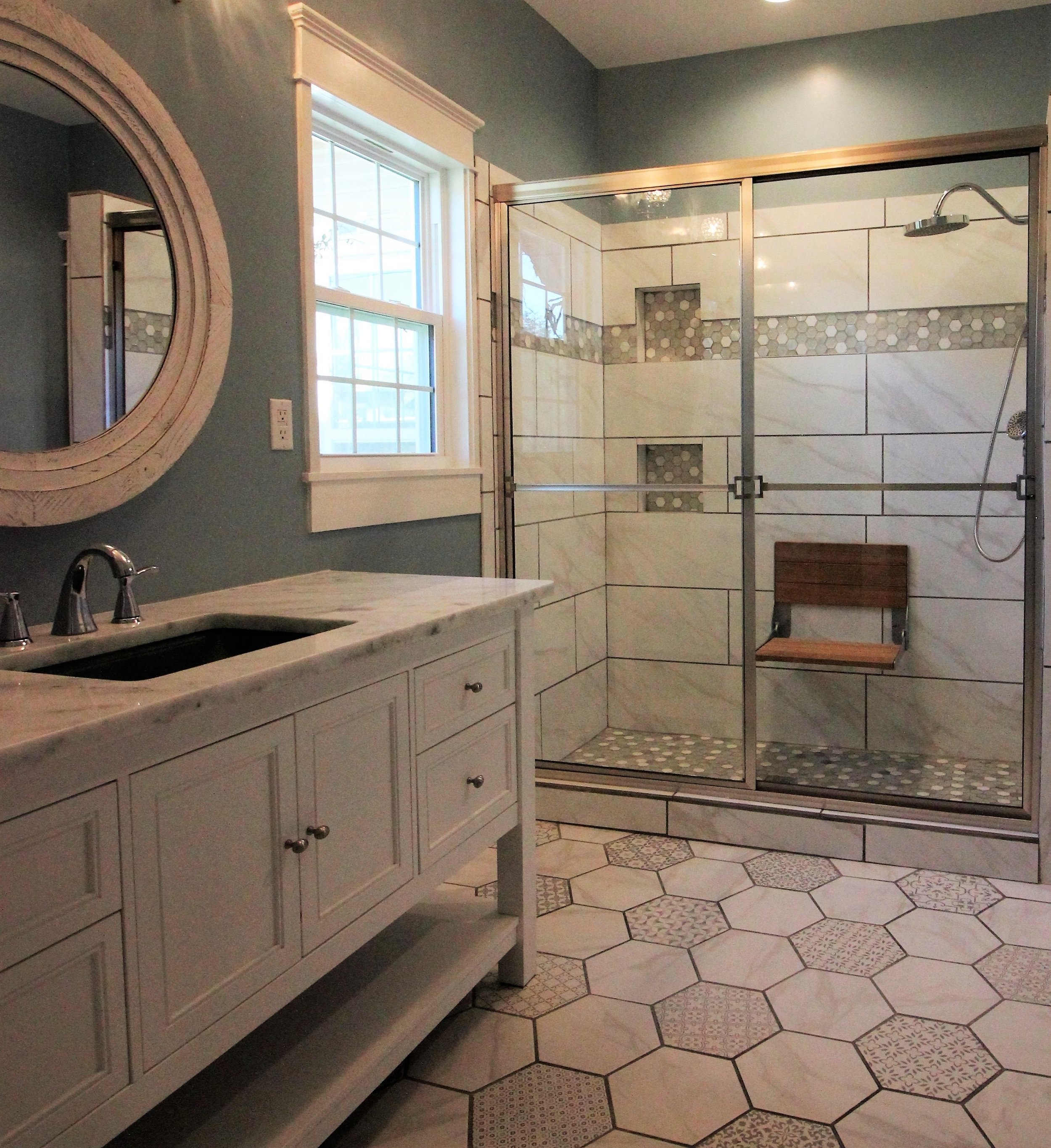 Bathroom with a white vanity, marble countertop, round mirror, window, and a glass shower enclosure with marble and hexagonal tile accents.