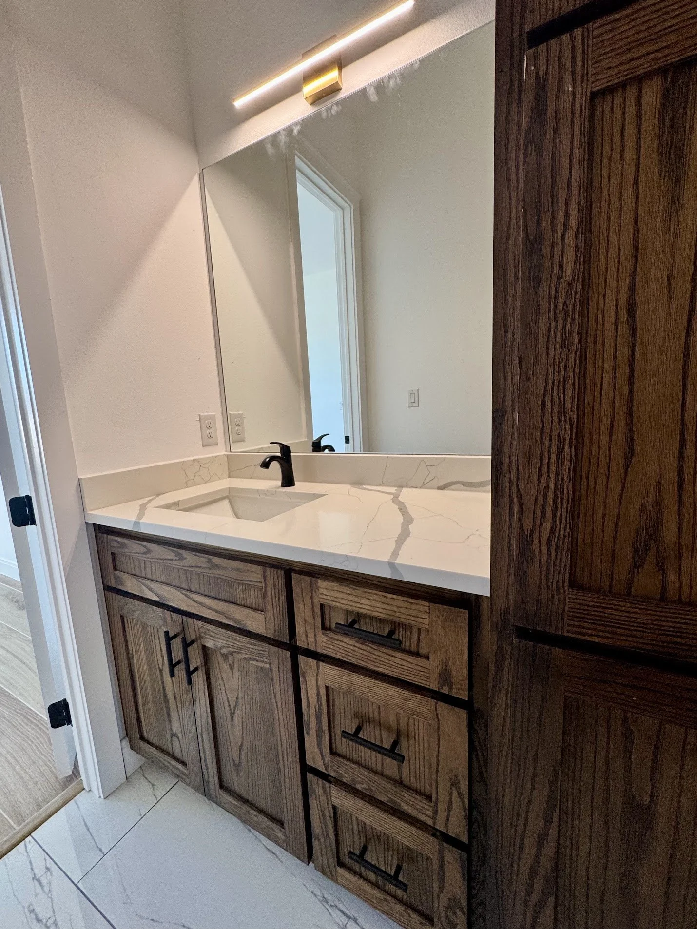 Bathroom vanity with a marble countertop, a black faucet, and wooden cabinets beneath, reflected in a large mirror. Soft lighting above the mirror.
