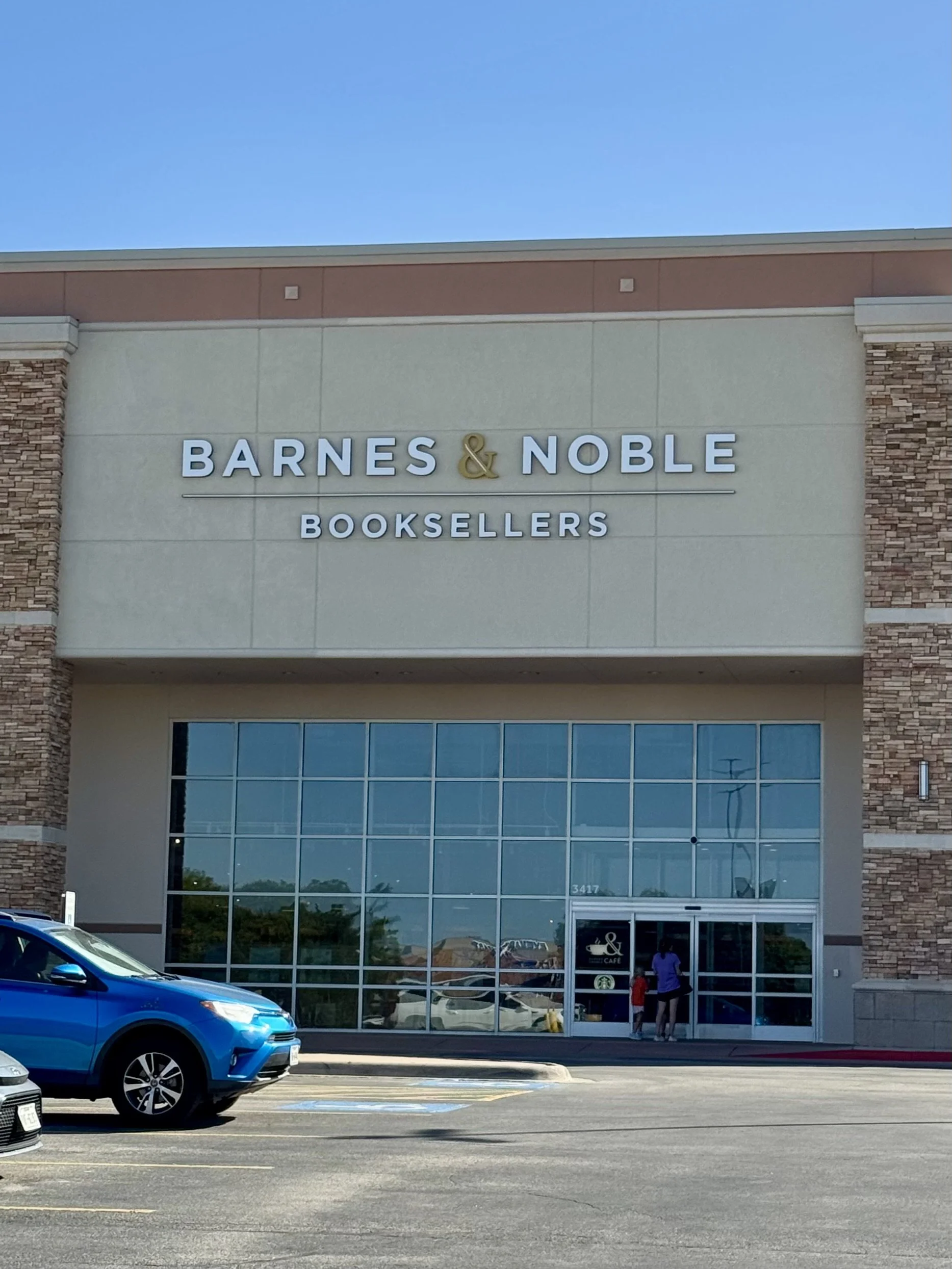 Outside view of Barnes & Noble bookstore with glass entrance, a parking lot with several cars, and a clear blue sky.