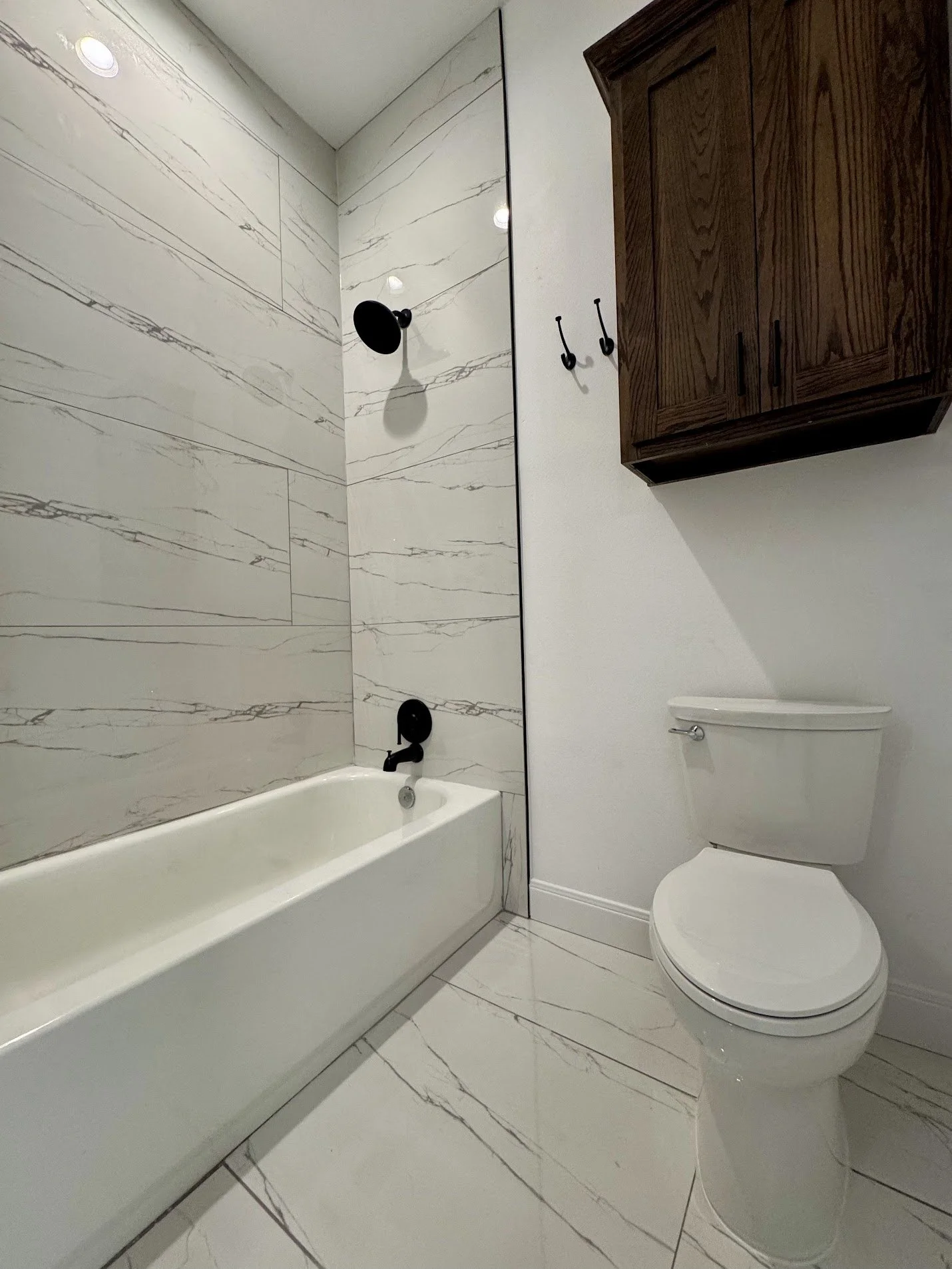 A modern bathroom with a white bathtub, black fixtures, a marble accent wall in the shower, a wall-mounted wooden cabinet, and a white toilet.