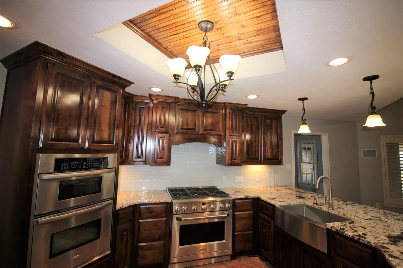 Kitchen with wooden cabinets, granite countertops, stainless steel oven and sink, white tiled backsplash, and hanging light fixtures.