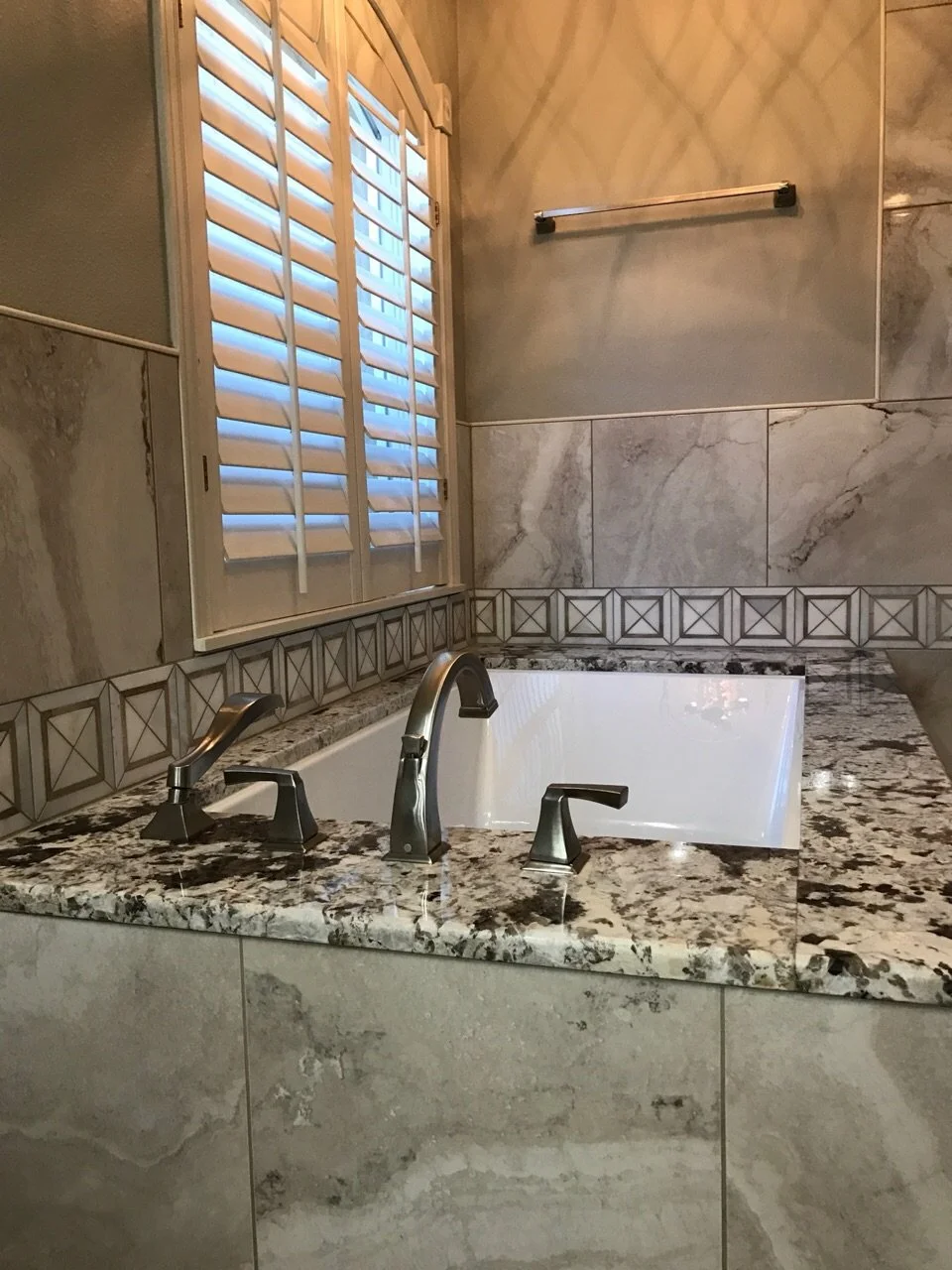 Bathroom with a built-in bathtub surrounded by a granite countertop, large marble tiles on the walls, a window with white plantation shutters, and a towel rack on the wall.