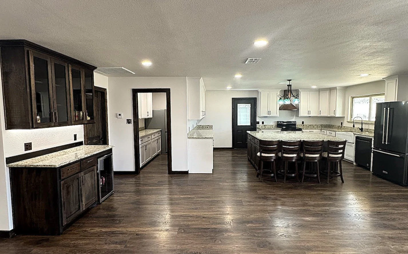 Kitchen with dark and white cabinets, granite countertops, hardwood floors, central island with chairs, black refrigerator, window above sink, and ceiling lights.