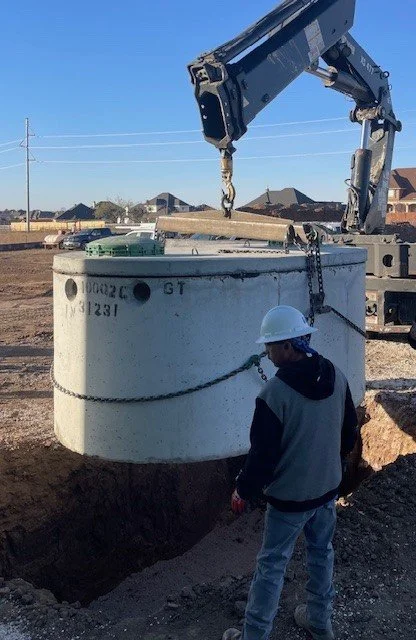 Construction site with worker in hard hat observing large concrete septic tank being lifted by crane.