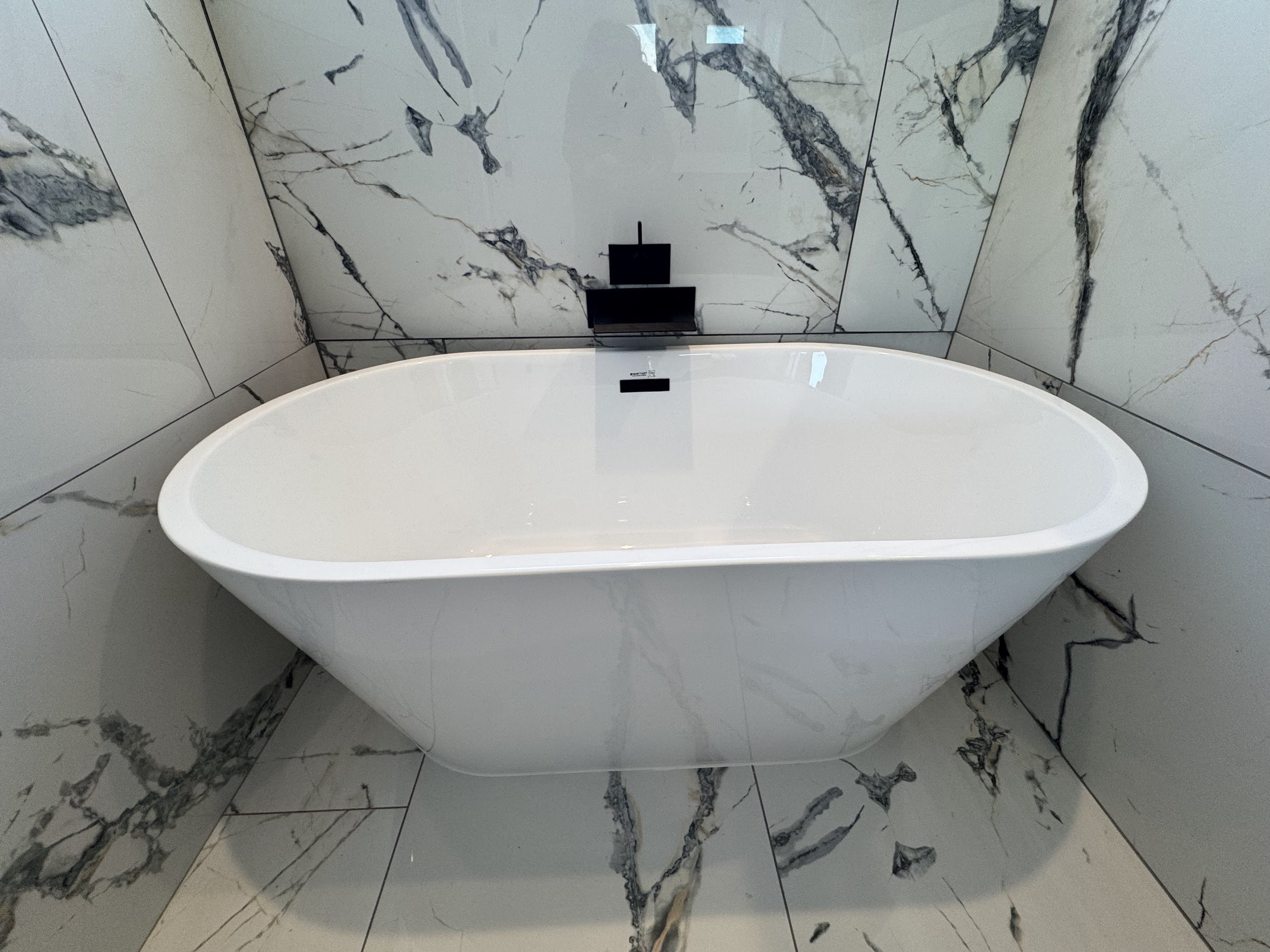 A modern white freestanding bathtub in a bathroom with marble walls and floor.