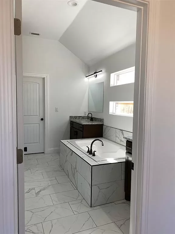 A modern bathroom with white marble tile flooring and a built-in bathtub with black fixtures. A vanity with a granite countertop and a mirror is seen in the corner, with a black light fixture above. Two horizontal windows provide natural light, and a white door is on the left side.