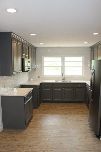 Empty modern kitchen with dark gray cabinets, white countertops, stainless steel appliances, a double window above the sink, and wood flooring.