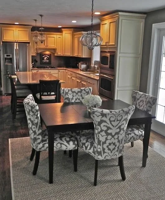 A kitchen and dining area with a black dining table surrounded by six patterned upholstered chairs. The kitchen has wooden cabinets, stainless steel appliances, and a central island. A light fixture hangs above the table, and there is a large window with natural light.