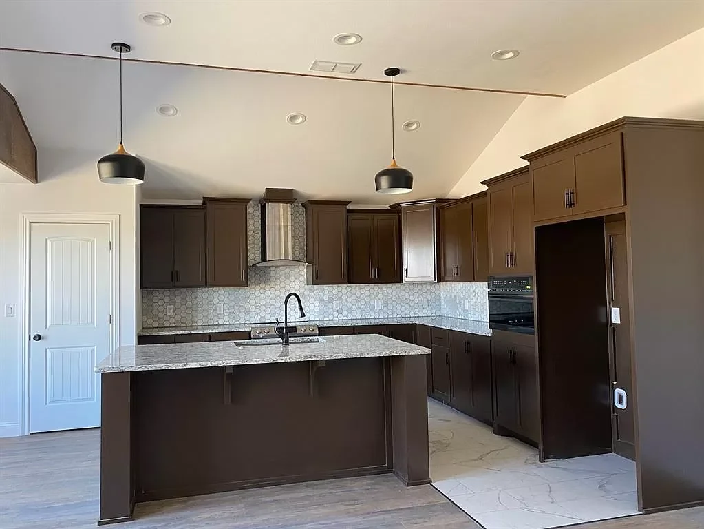 Modern kitchen with dark brown cabinets, a granite island, black pendant lights, light-colored backsplash, and built-in appliances.
