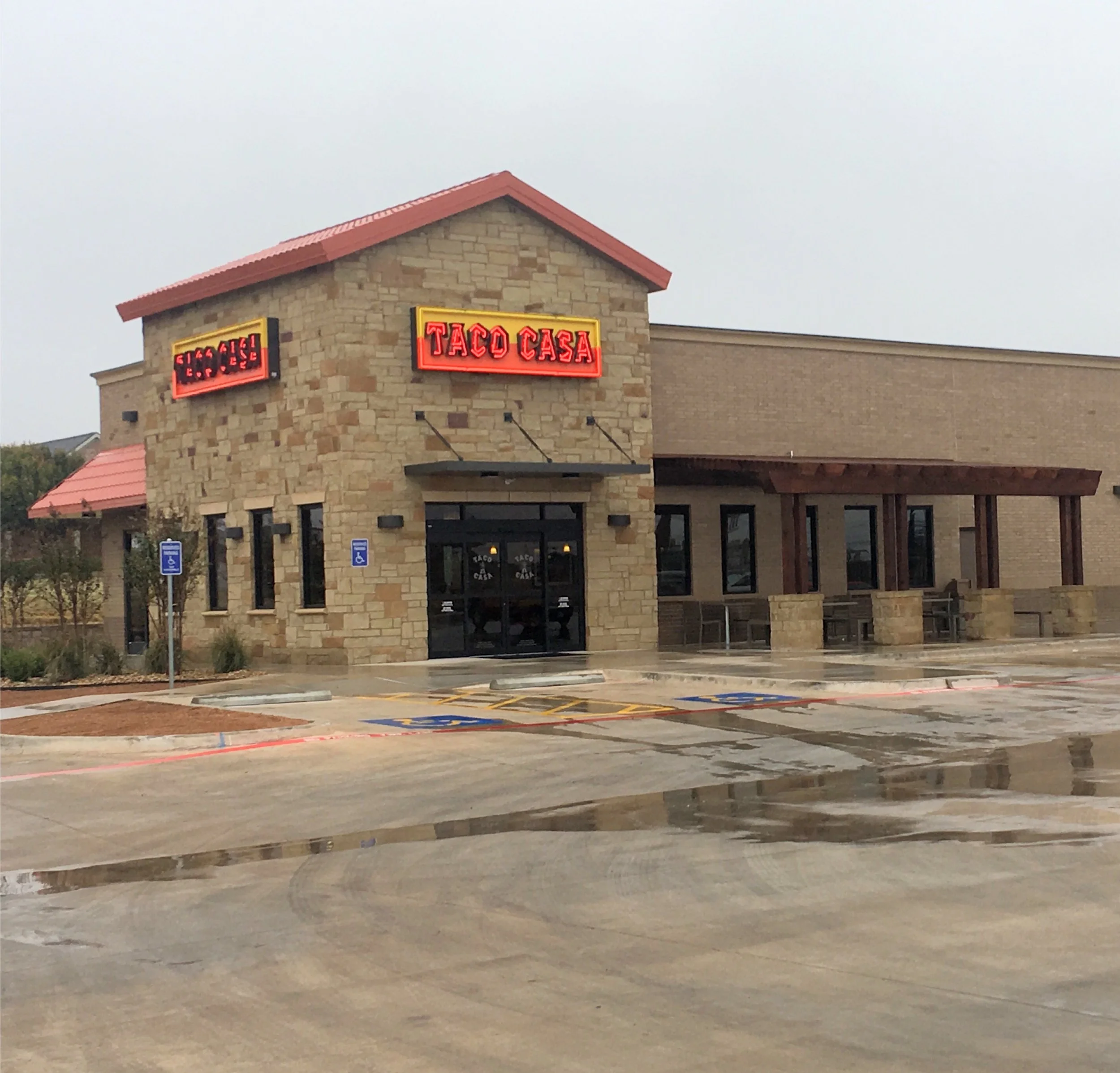 Exterior of a Taco Casa fast-food restaurant with stone facade, red signage, and accessible parking spaces in front on a wet day.