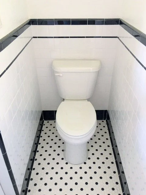 A small bathroom with a white toilet, white tiled walls with black trim, and black and white mosaic floor tiles.
