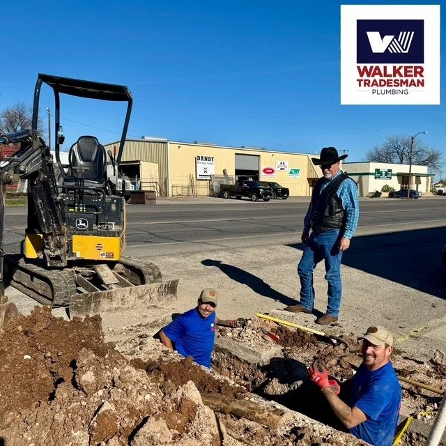 Three men working on a sidewalk construction project on a city street, with a small excavator and buildings in the background, one wearing a cowboy hat.