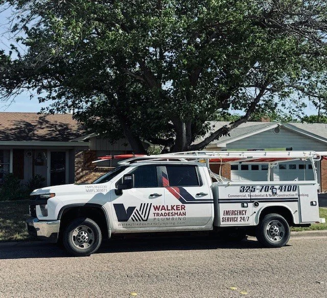 A white plumbing service truck parked on a residential street with a large tree and houses in the background. The truck has company branding for Walker Tradesman Plumbing, including contact information and service details.