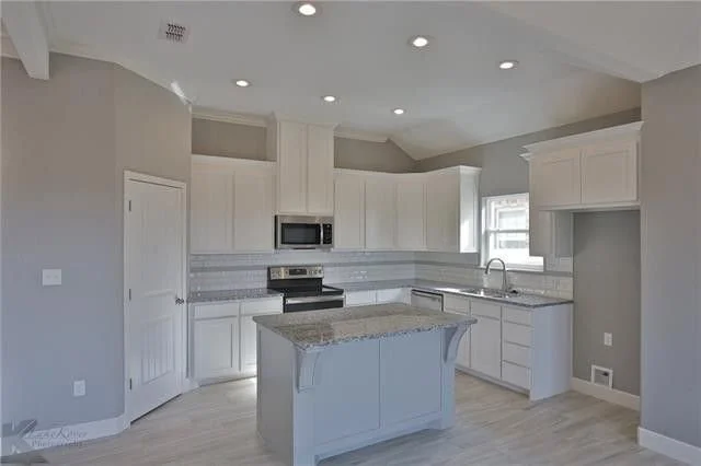 Empty modern kitchen with white cabinets, a small island with a granite countertop, stainless steel stove and microwave, sink under a window, and light gray walls.