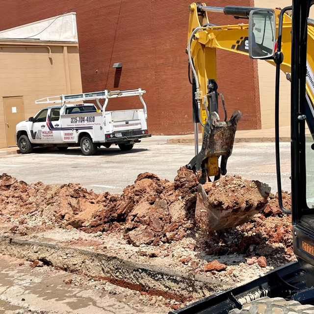 View of construction site with a yellow backhoe digging into the ground, large dirt and rocks in the foreground, and a white utility truck parked near a brick building in the background.