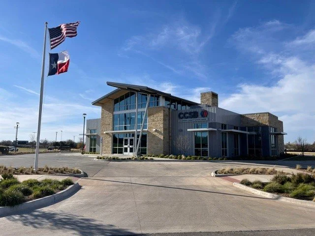 A modern building with large glass windows and a brick exterior, with an American flag and a Texas state flag flying on flagpoles in front of it, under a blue sky.