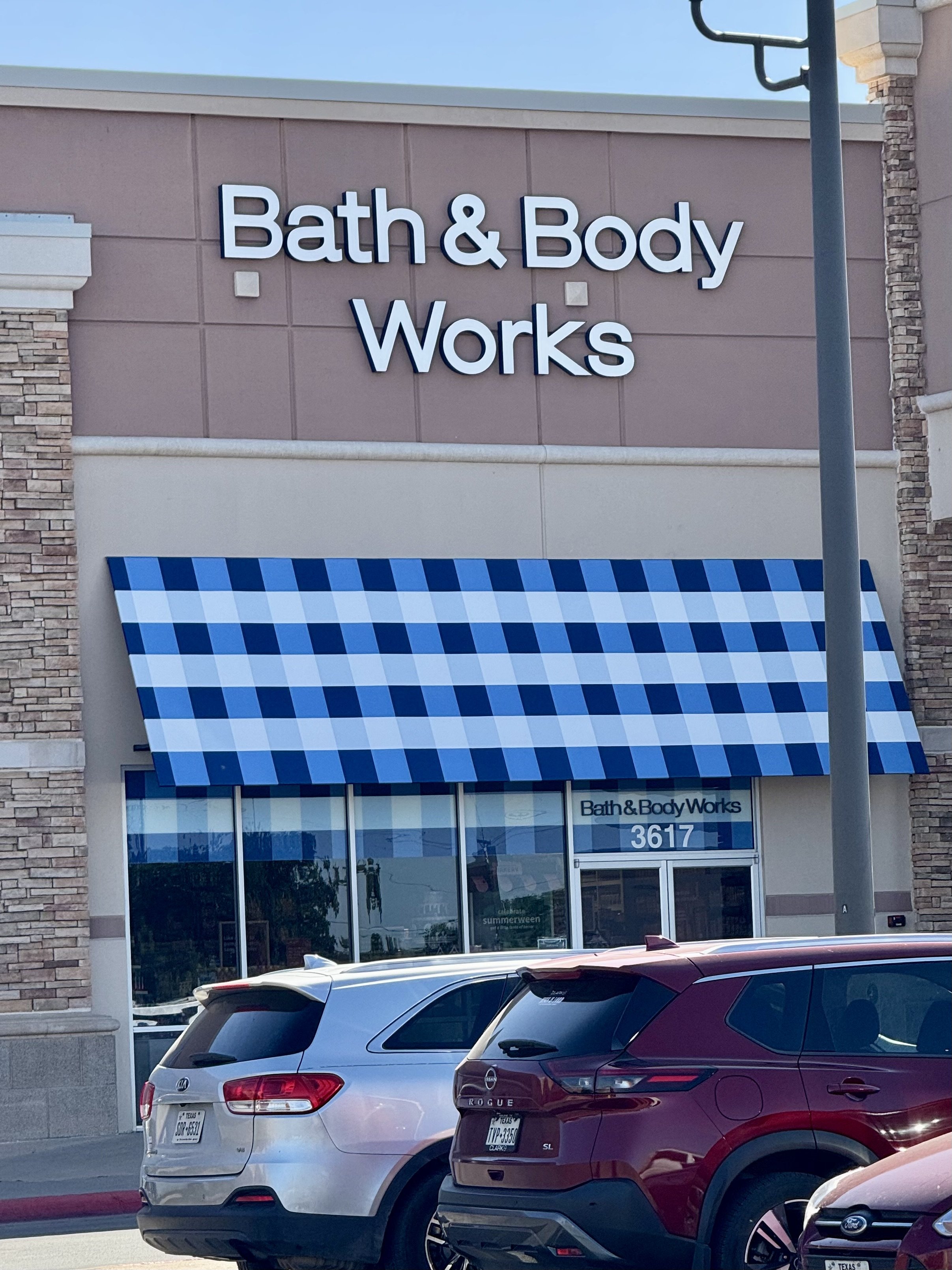 Exterior view of a Bath & Body Works store with a blue and white checkered awning, parked cars in front, and the store's sign on the building.