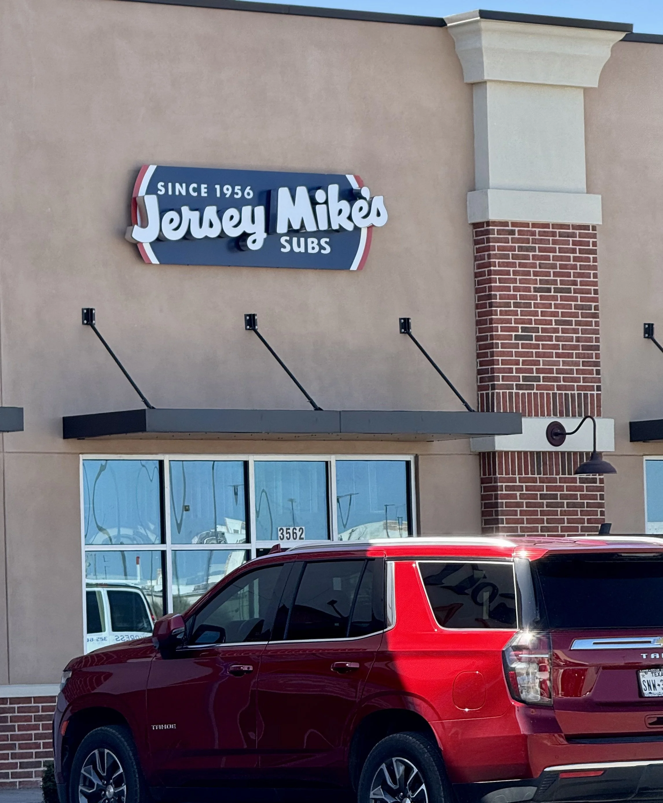 Jersey Mike's sandwich shop with a parking lot in front, a red SUV parked near the window, and a sign that says 'Since 1956 Jersey Mike's Subs'.