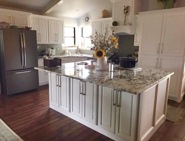 Modern kitchen with white cabinets, granite island, stainless steel refrigerator, and wooden flooring, decorated with flowers on the island.