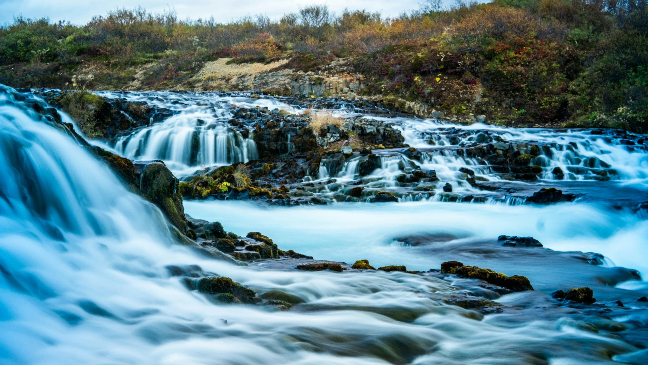 A scenic waterfall flowing over rocks with a wooded hillside in the background.