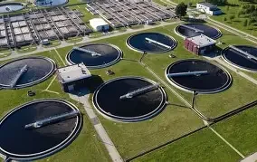 Aerial view of a wastewater treatment plant with multiple circular clarifiers and processing tanks surrounded by green grass and pathways.