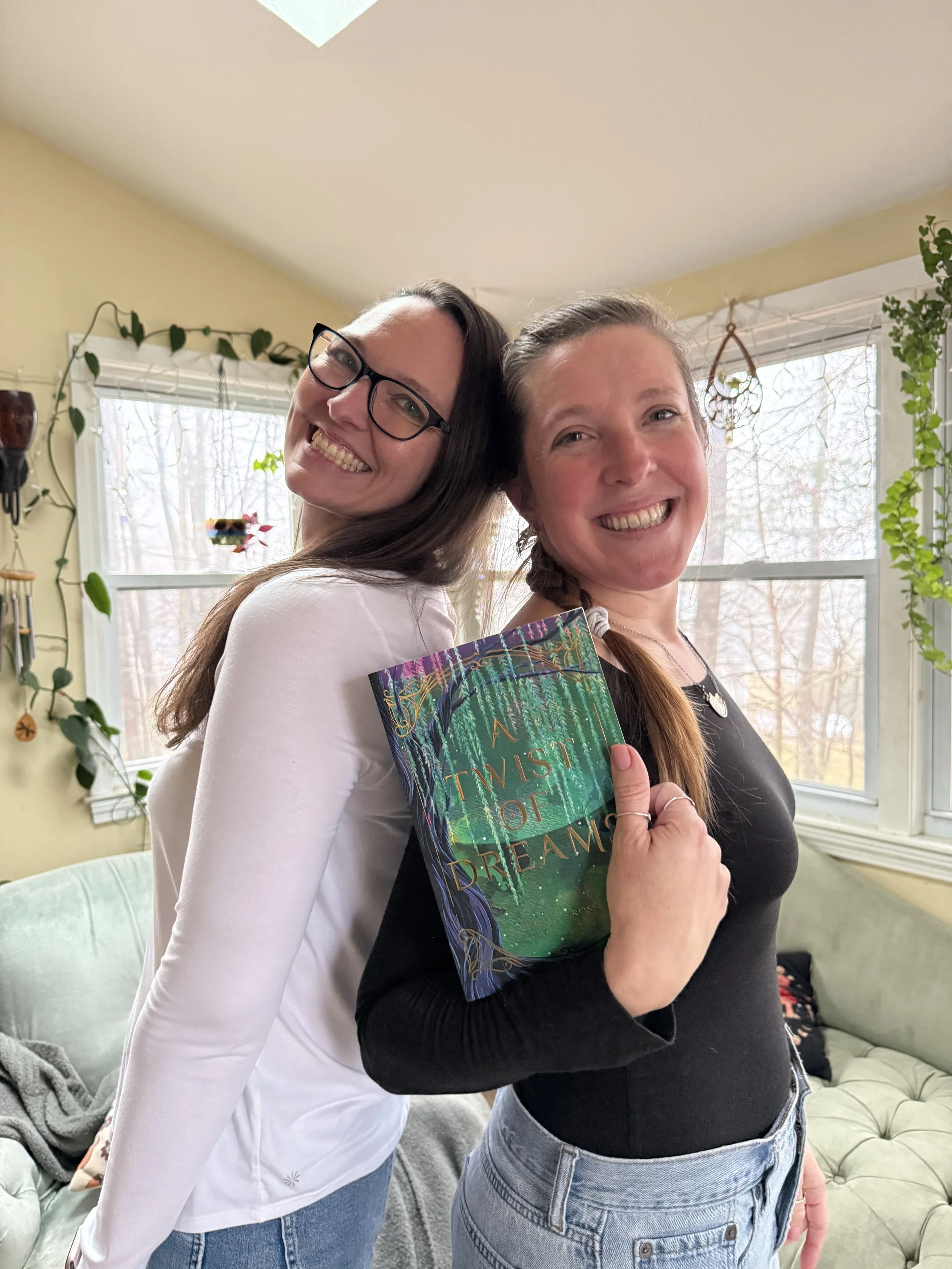 Two women smiling and leaning against each other in a cozy, plant-filled living room. One woman is holding a book titled 'A Twist of Dream.'