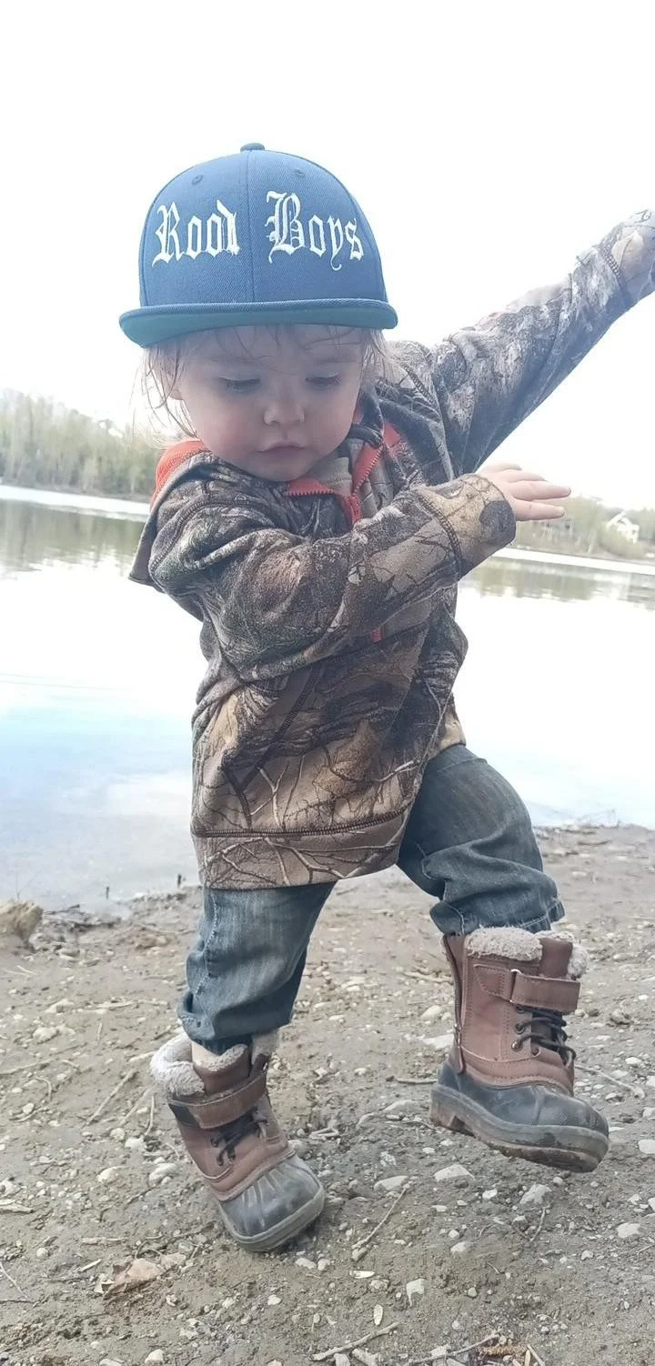 Young child standing on rocky ground near water, wearing a camouflage jacket, jeans, brown boots, and a blue cap