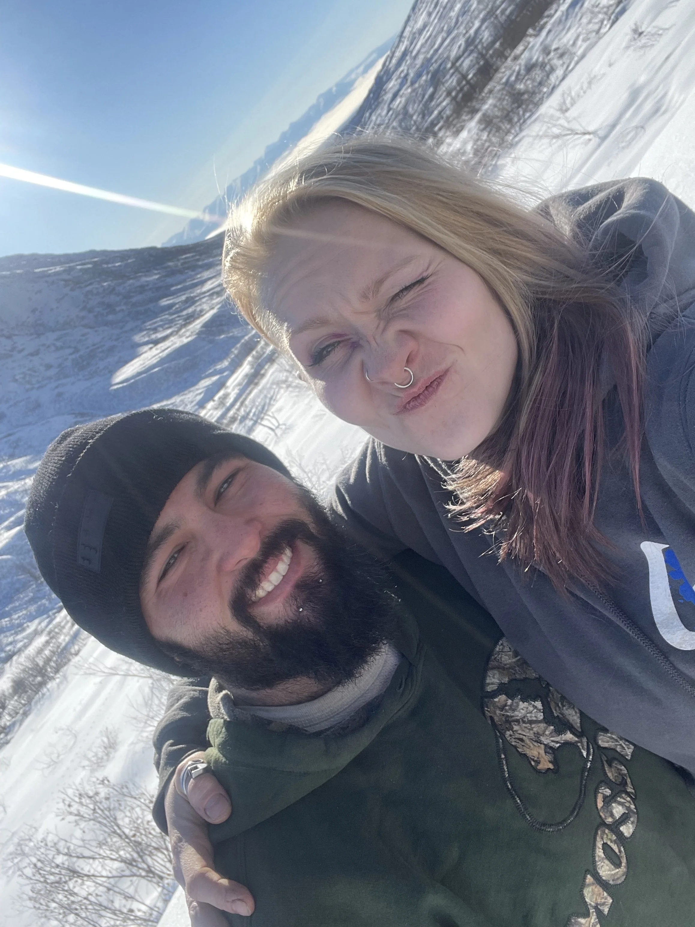 Two people outdoors in a snowy landscape, one with blonde hair making a squinting face and wearing a nose ring, the other with a beard and wearing a black beanie, smiling. They are dressed warmly and seem to be enjoying a winter day.