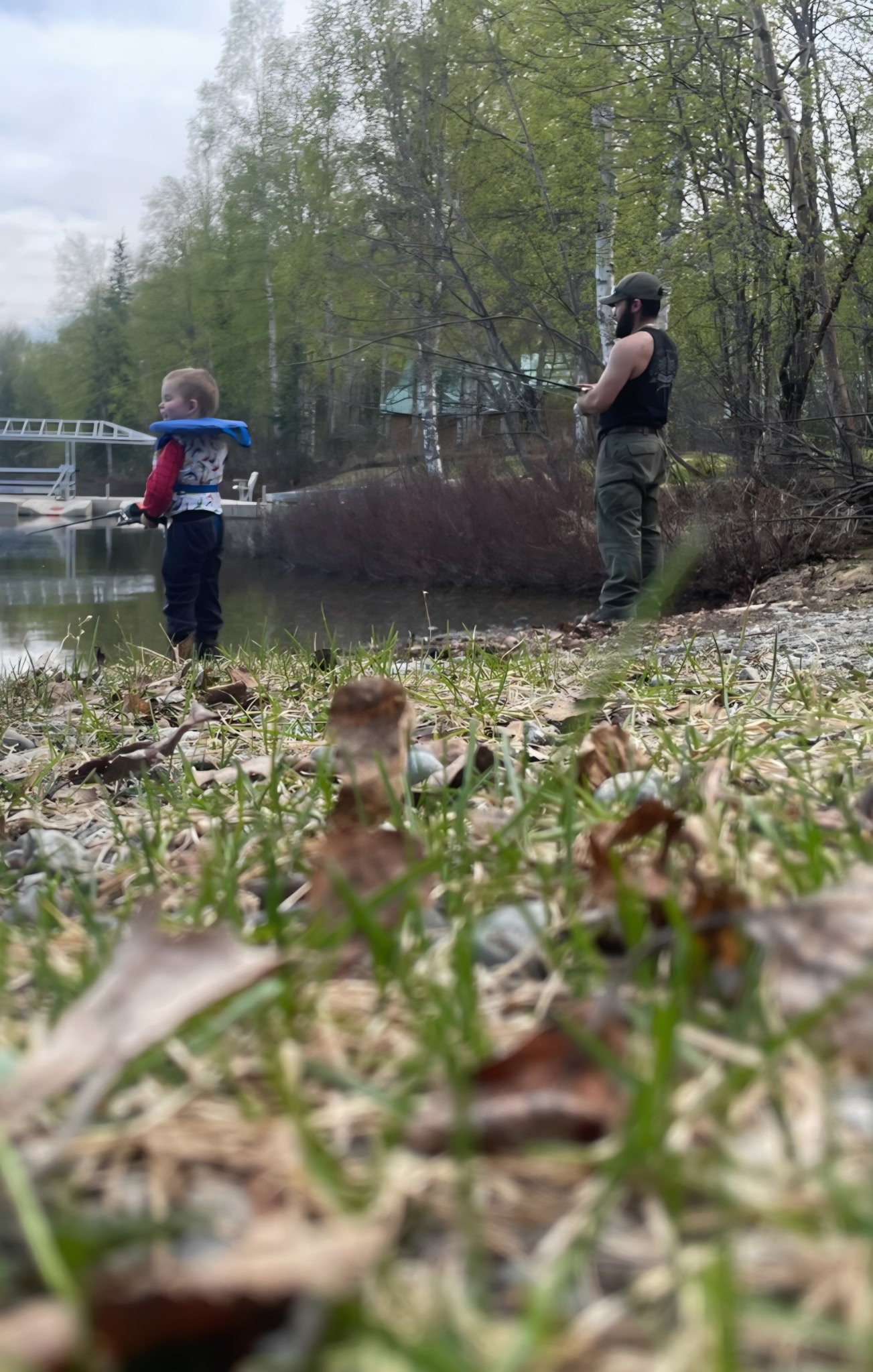 A person wearing a hat and sleeveless shirt fishing by a wooded lakeshore with a child in a life jacket nearby fishing