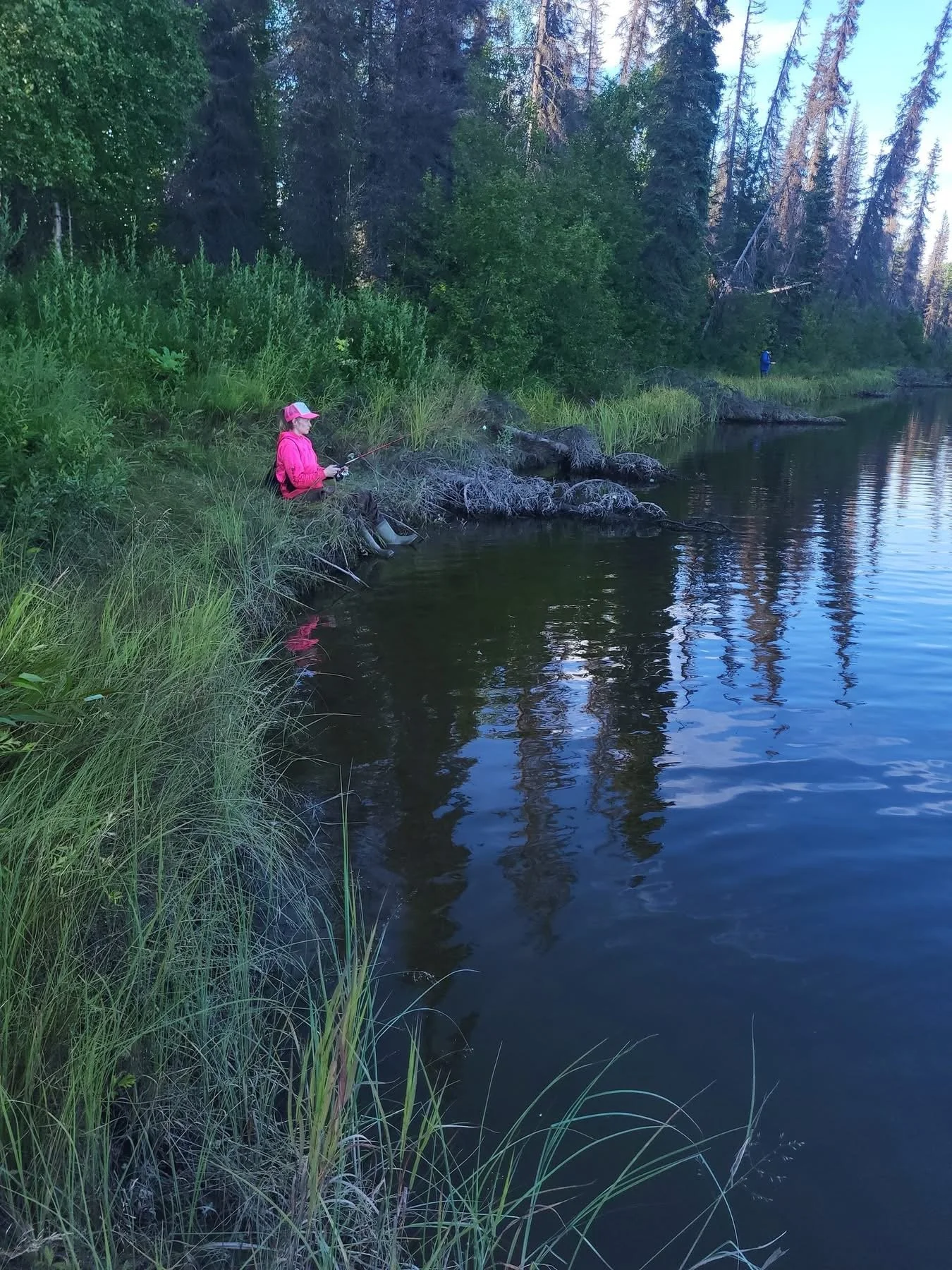 A woman in a pink jacket and cap is fishing by a river surrounded by lush green vegetation and tall trees.