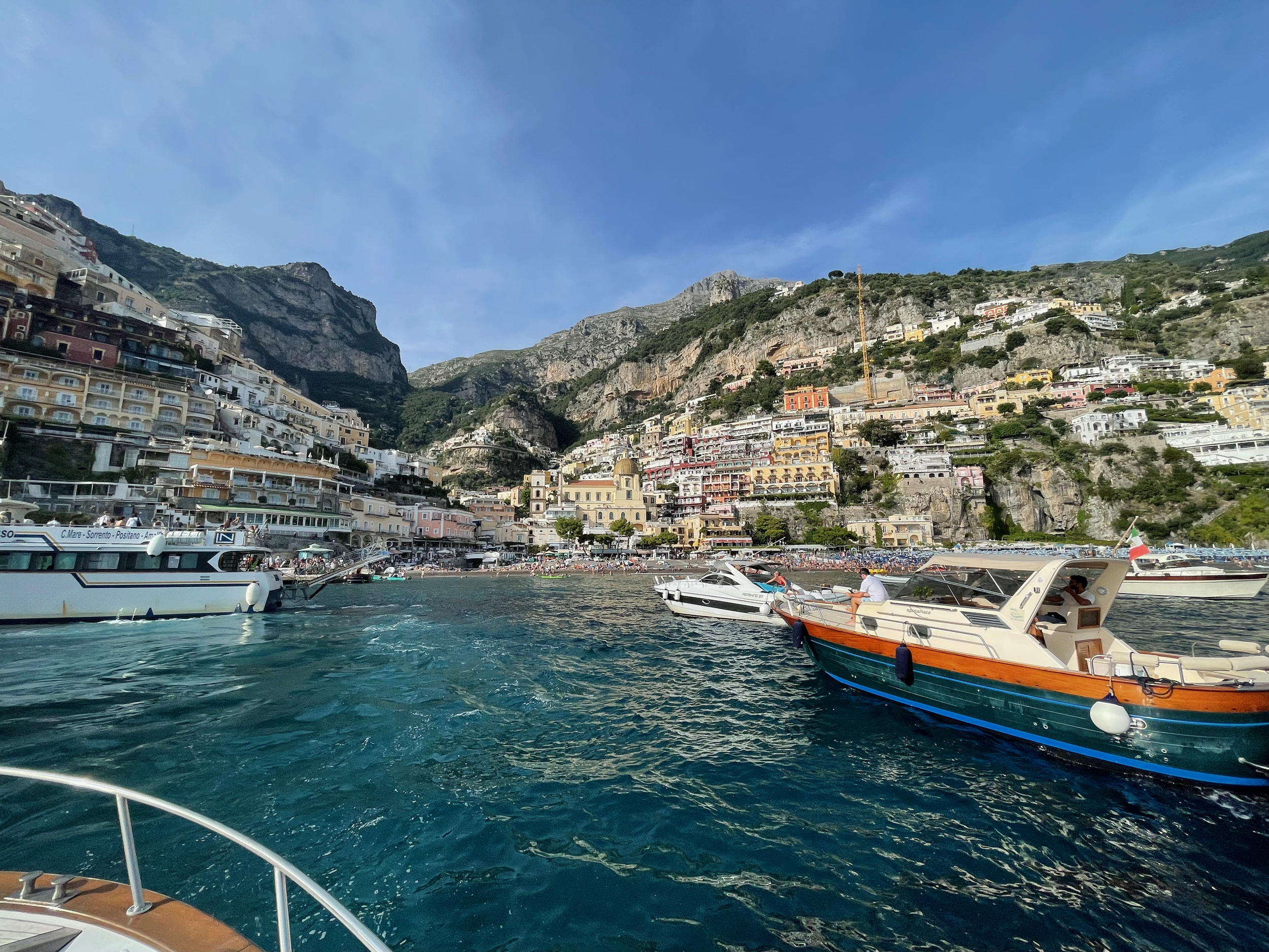 A harbor with boats docked in the water, with hillside buildings and mountainous terrain in the background under a blue sky.