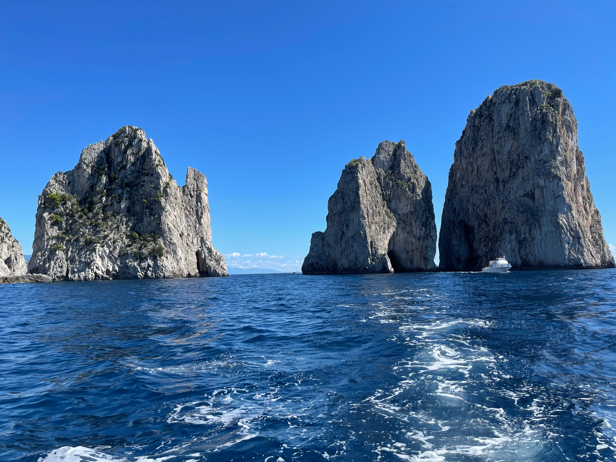 Large sea stacks rising from the ocean, with a bright blue sky overhead and a boat sailing nearby, from Capri, Italy.