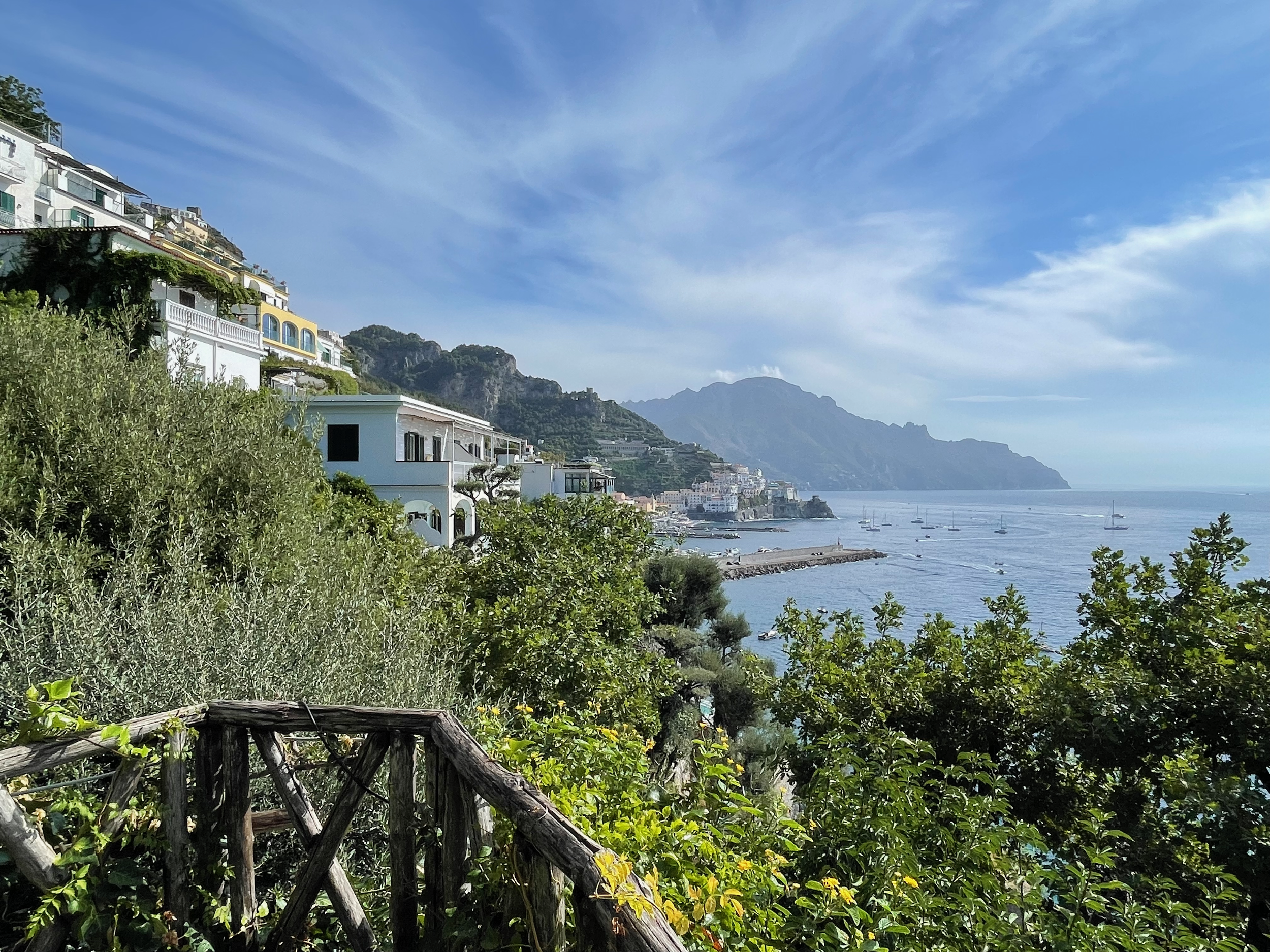 Scenic view of a coastal hillside with white and yellow buildings overlooking the ocean, with sailboats, lush greenery, and mountains in the background under partly cloudy sky.