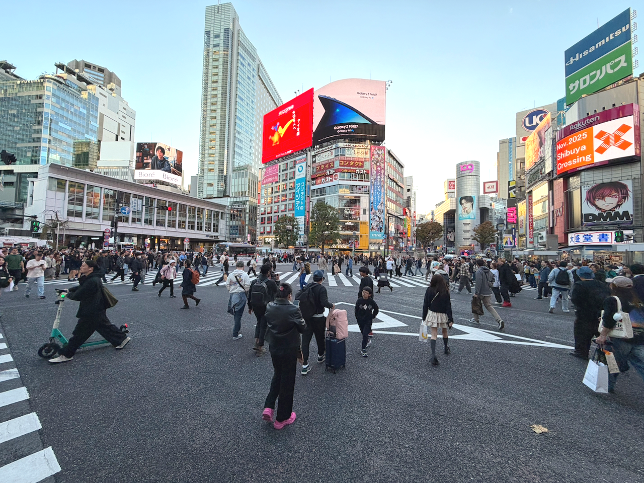 Crowded intersection in Tokyo's Shibuya district with many pedestrians crossing the street. Tall buildings with colorful billboards and electronic screens surround the area.