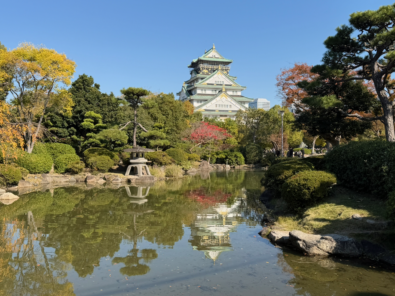 A traditional Japanese castle with multiple tiers and ornate roofs, surrounded by a lush garden with trees, shrubs, a pond, and a stone lantern, under a clear blue sky.